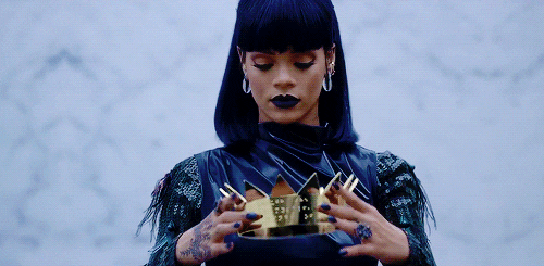 Rihanna de cabelo preto, franja e roupa preta, levantando uma coroa e colocando na cabeça 