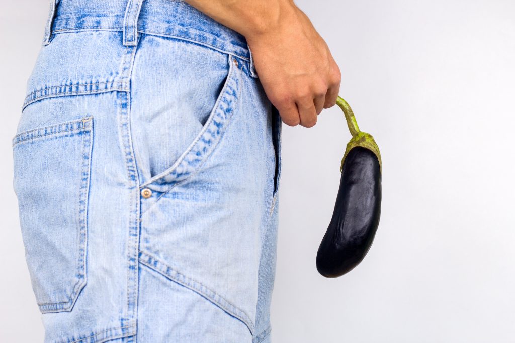 Homem de calça jeans segurando uma beringela na frente do penis para remeter a disfunção erétil