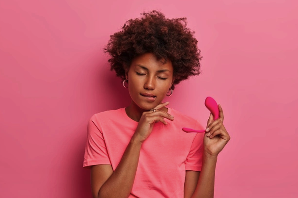 mulher em um fundo rosa com os olhos fechado e a língua entre os dentes demonstrando prazer. Ela segura um vibrador em sua mão esquerda.