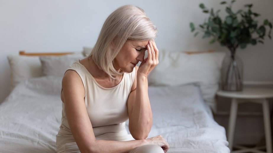 Senhora sentada na cama com a cabeça abaixada e a mão na cabeça, frustrada devido a secura vaginal na menopausa