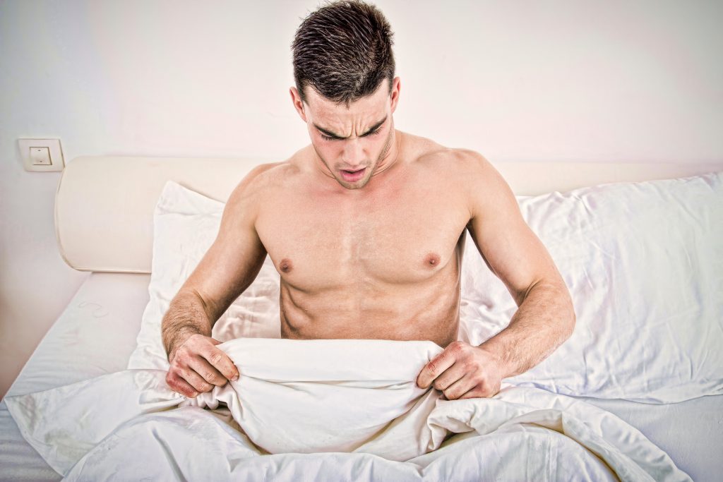 Homem sem blusa deitado na cama, levantando o edredom e olhando para baixo em direção ao seu órgão sexual