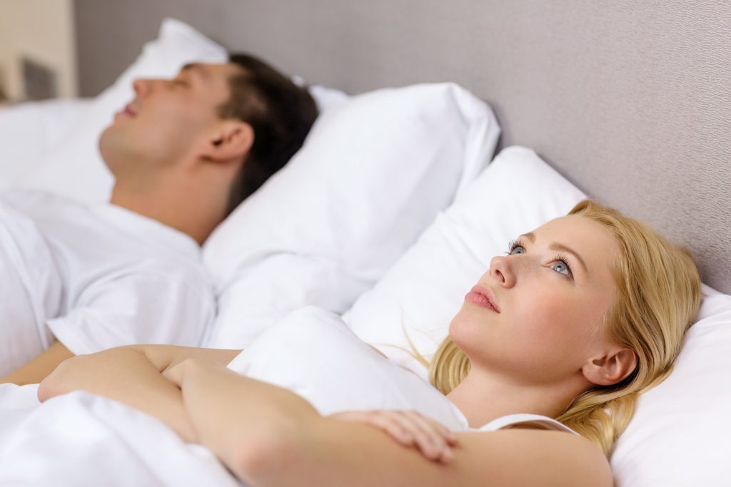 Homem de camiseta branca, dormindo em cama branca, ao lado de uma mulher loira de olhos azuis, com cara de insatisfeita ao lado 