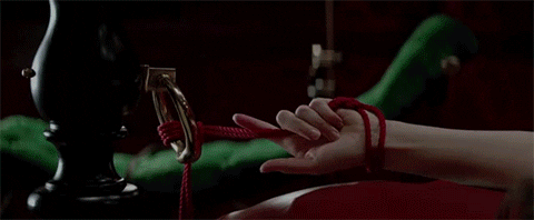 Mulher vendada, com a boca aberta, deitada em sofá vermelho, amarrada com uma corda vermelha na cama. Cena do filme 50 tons de cinza 