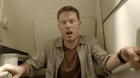 Homem com camiseta marrom e camisa, sentado na cabine de um avião, de olhos fechados, boca aberta, tendo um orgasmo
