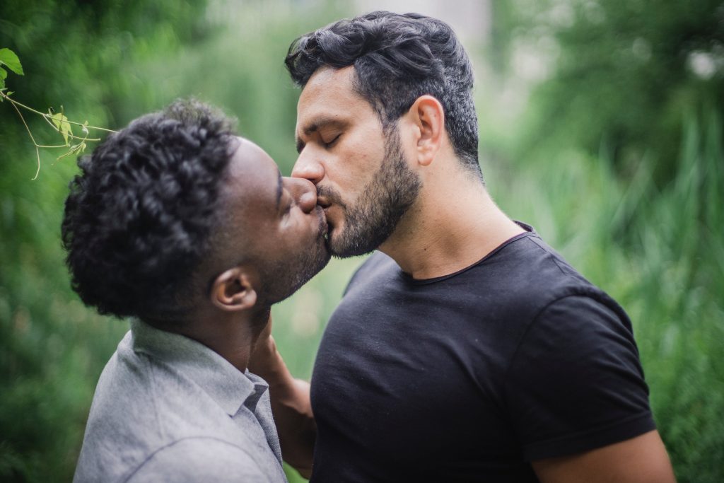Homem de camisa branca beijando outro homem de camisa preta, em fundo desfocado de natureza