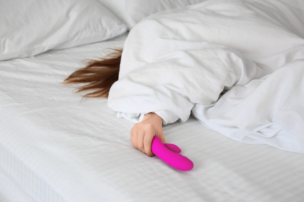 Mulher embaixo de um edredom branco com a mão pra fora segurando um vibrador rabbit ponto G cor rosa a fim de praticar a masturbação feminina