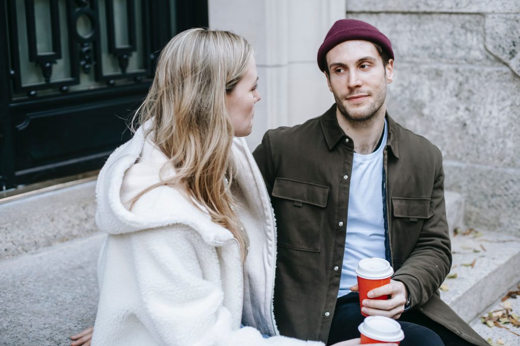 Homem e mulher no frio se olhando sentados em um banco tomando café 