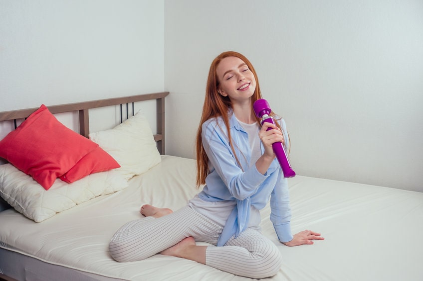 mulher feliz sorrindo sentada na cama com um vibrador na mão como se fosse um microfone