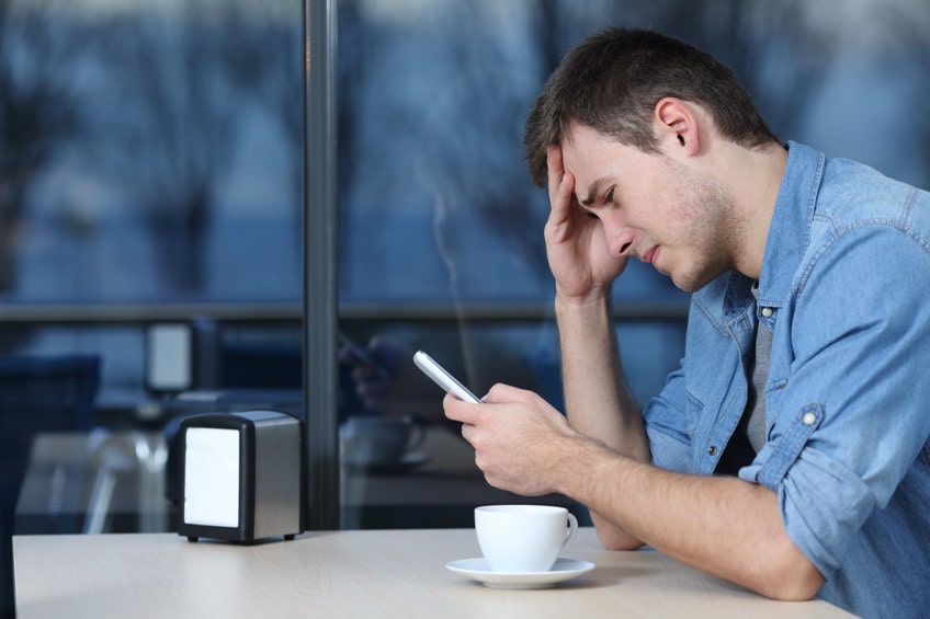 Homem sentado em uma mesa tomando café, chorando olhando o celular, sendo vítima de ghosting