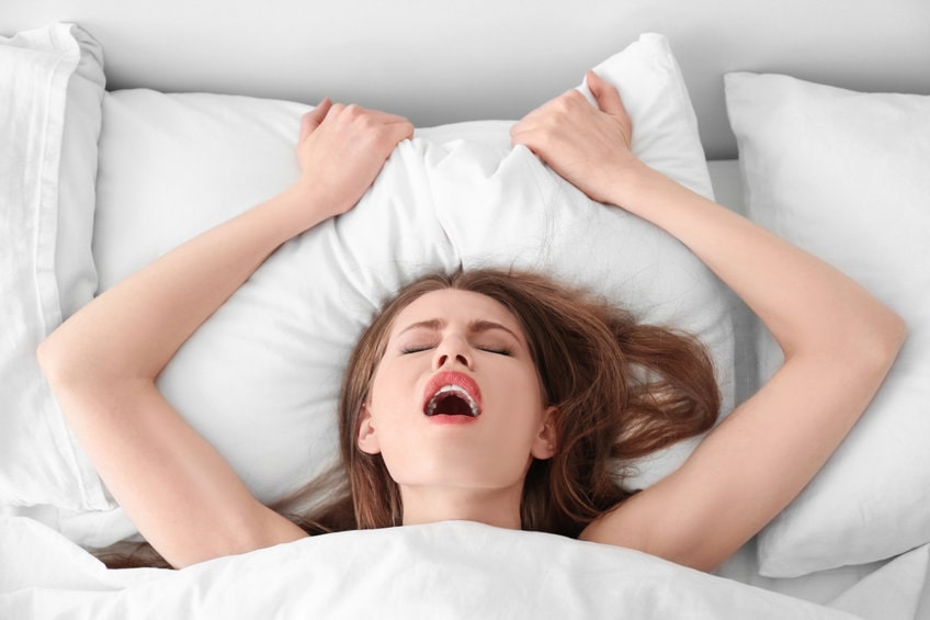 mulher deitada de baixo do edredom, com os olhos fechados, boca aberta segurando o travesseiro com as duas mãos, tendo orgasmos múltiplos