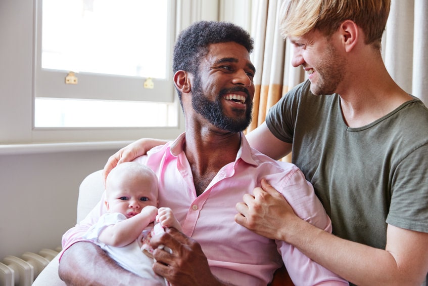 orgulho lgbtqia+: homem negro de camisa rosa sorrindo e segurando um bebê no colo, ao lado de um homem loiro de camiseta verde o abraçando e sorrindo 