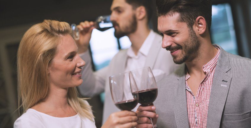 mulher e homem se olhando e sorrindo, brindando com duas taças de vinho, enquanto o outro observa atrás, praticando cuckold