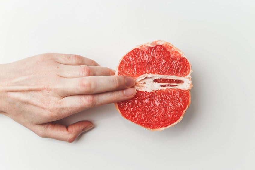 Mão tocando uma laranja vermelha, simulando o toque no clitóris