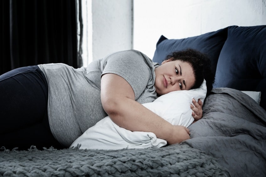 Mulher de camiseta cinza deitada no sofá abraçando o travesseiro, triste devido a anorgasmia